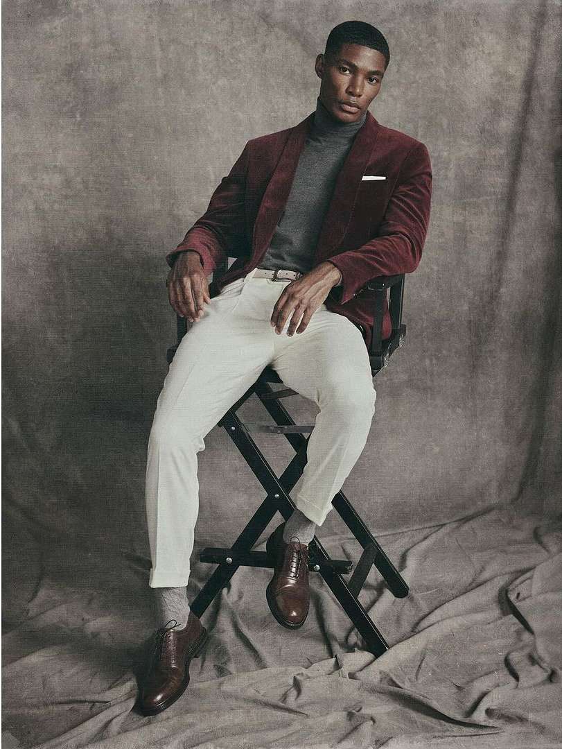 Portrait of a handsome African-American man in a red jacket sitting on a director's chair against a textured grey backdrop in a studio.