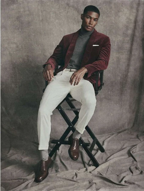 Portrait of a handsome African-American man in a red jacket sitting on a director's chair against a textured grey backdrop in a studio.