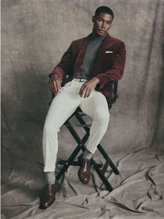 Portrait of a handsome African-American man in a red jacket sitting on a director's chair against a textured grey backdrop in a studio.