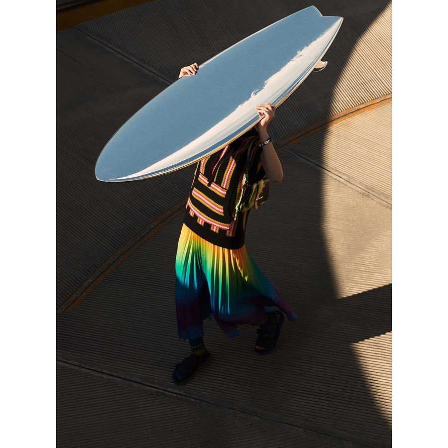 Person holding a surfboard with a colorful skirt and striped shirt standing on a textured grey surface in outdoor setting.