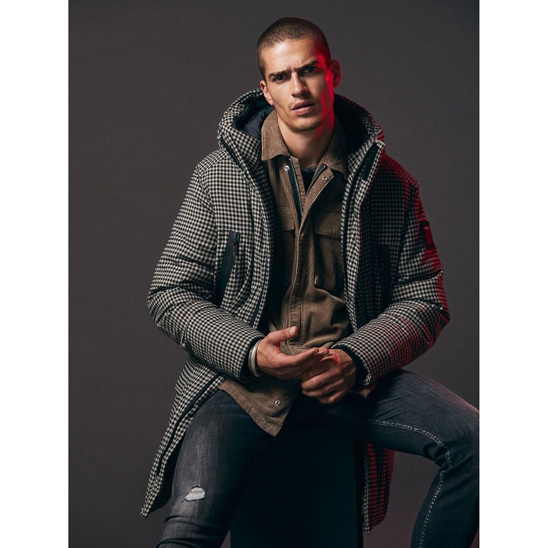 Portrait of a light-skinned adult male in a grey patterned hooded coat and dark jeans sitting in a photo studio against a grey backdrop.