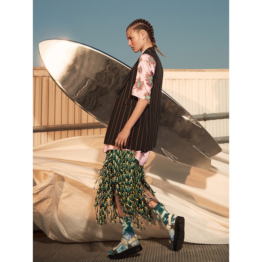 Fashion shot of a woman wearing a pinstripe vest, printed top, beaded skirt, platform sandals, and socks, standing next to a surfboard.