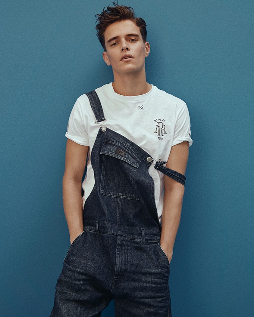A young man with brown hair poses in a white T-shirt and dark denim overalls against a solid blue background, hands in pockets.