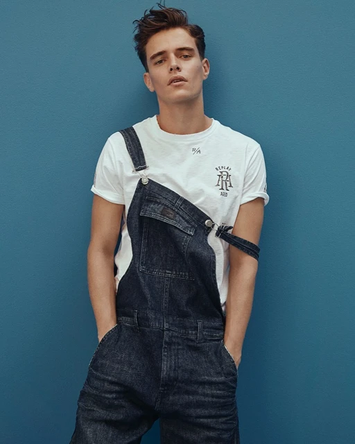 A young man with brown hair poses in a white T-shirt and dark denim overalls against a solid blue background, hands in pockets.