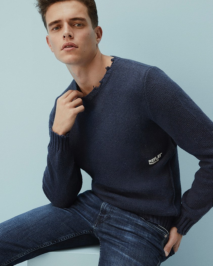 A seated male model wearing a dark blue knit sweater and denim jeans against a light blue backdrop.
