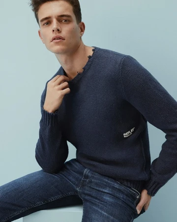 A seated male model wearing a dark blue knit sweater and denim jeans against a light blue backdrop.