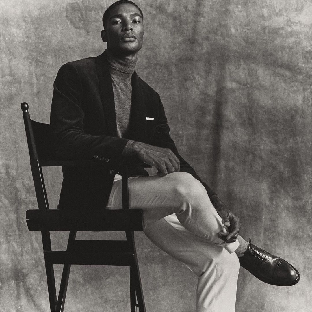 Elegant African American man in a velvet blazer and grey turtleneck, sitting on a black chair against a mottled grey backdrop in studio.
