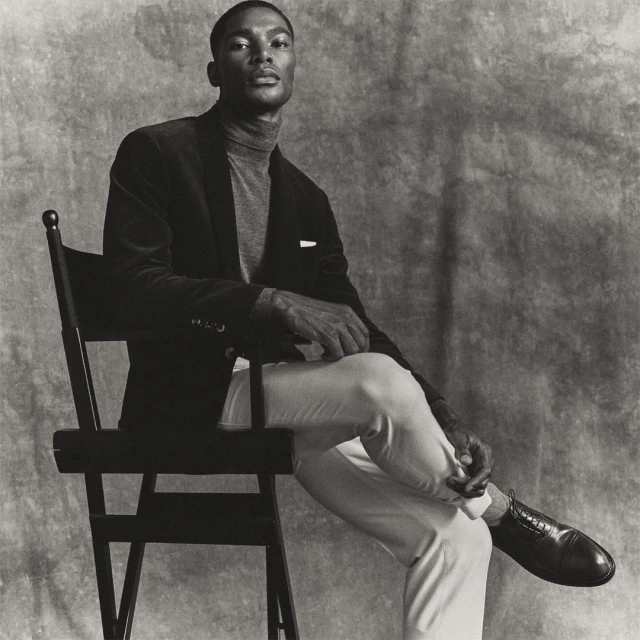 Elegant African American man in a velvet blazer and grey turtleneck, sitting on a black chair against a mottled grey backdrop in studio.