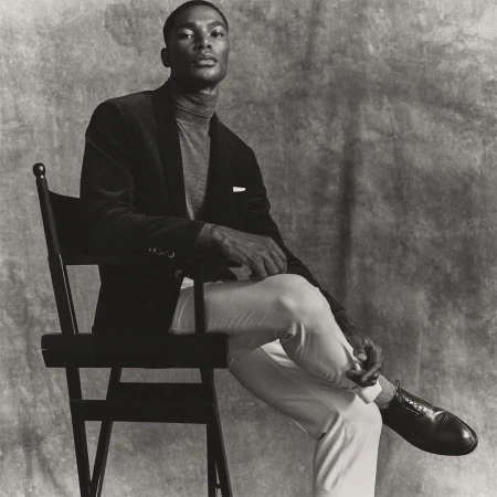 Elegant African American man in a velvet blazer and grey turtleneck, sitting on a black chair against a mottled grey backdrop in studio.