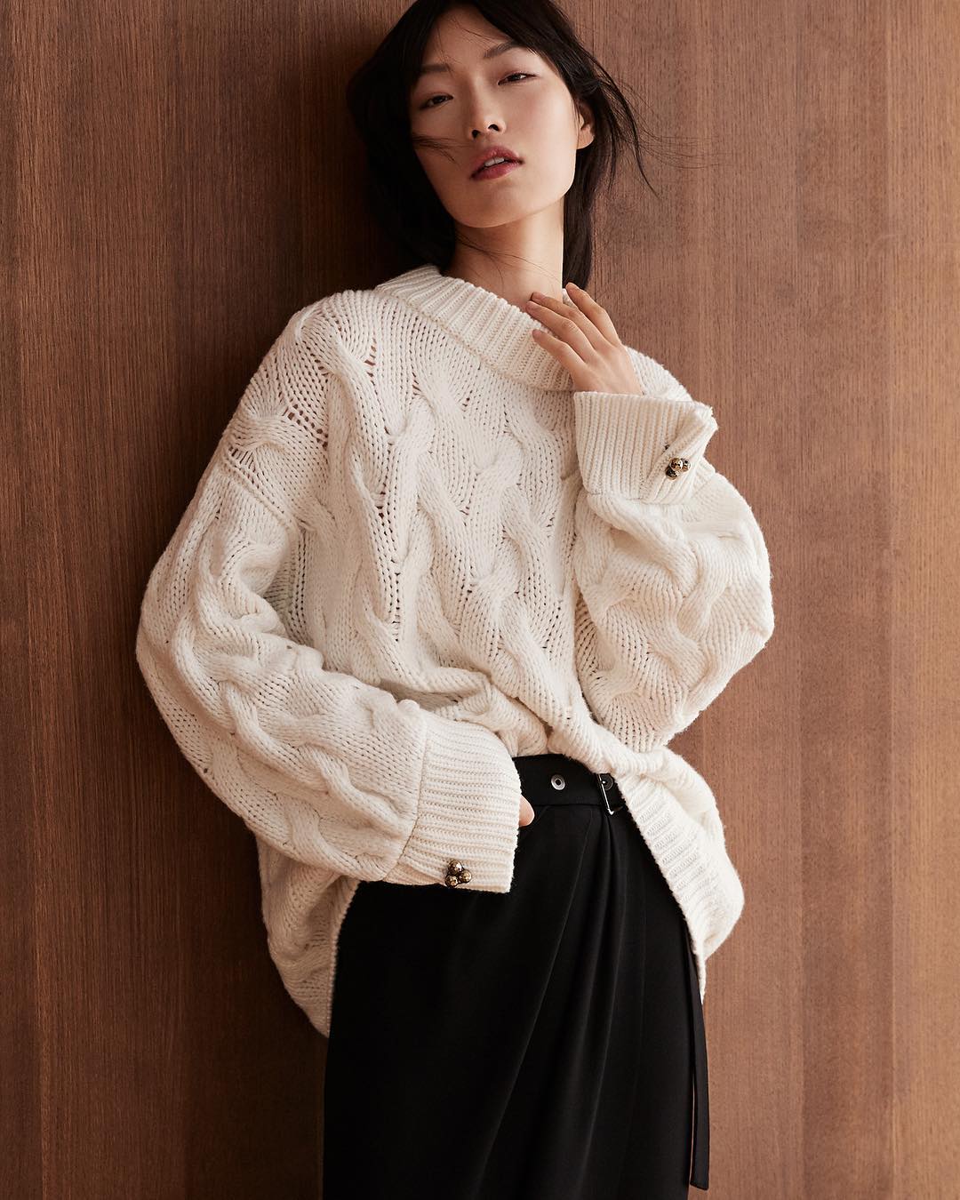 Portrait of an Asian woman wearing an ivory-colored, cable-knit sweater and black skirt, leaning against a wood wall indoors.