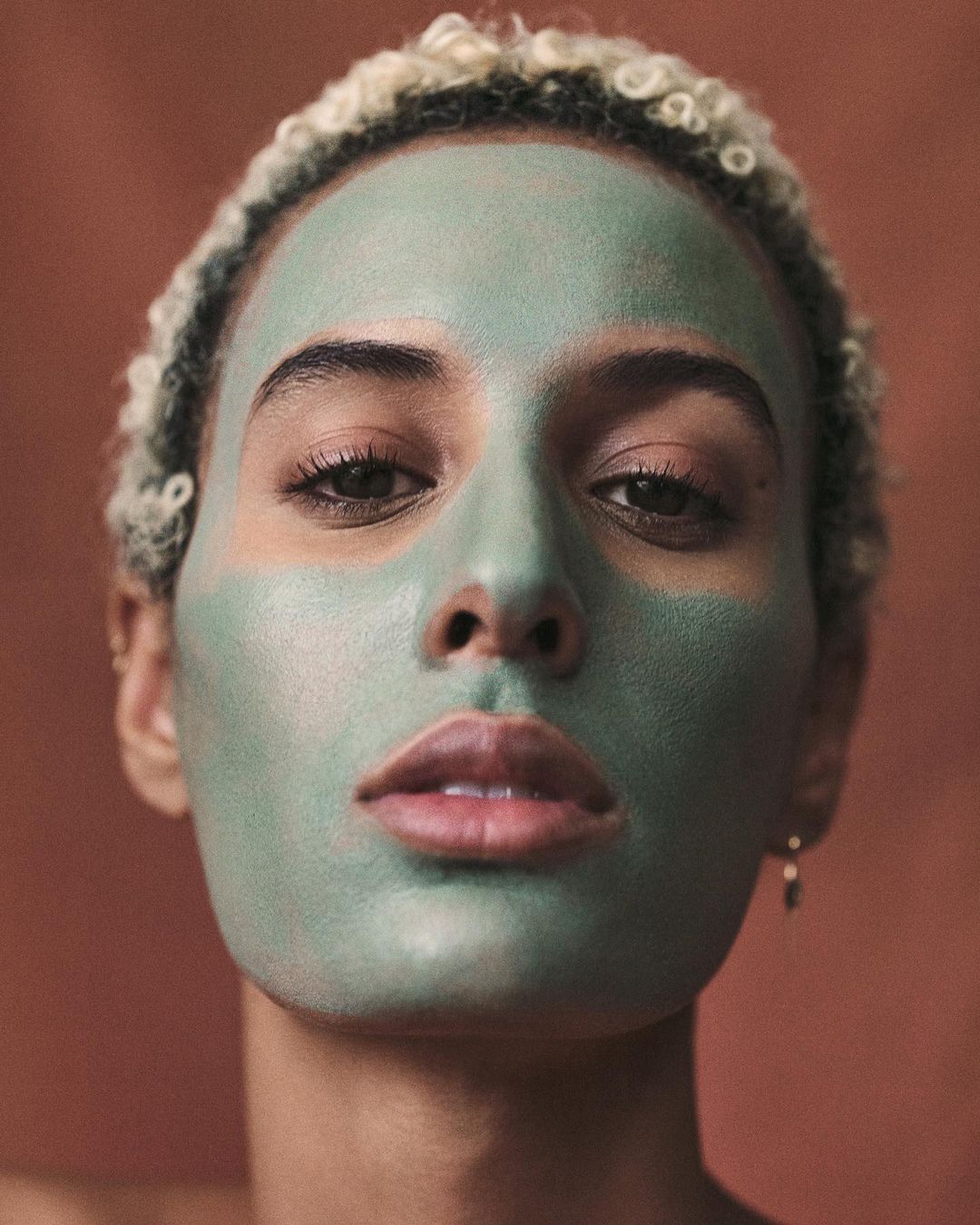 Close-up portrait of a young adult female with a green facial mask, fair skin, and light hair against a muted reddish-brown background.