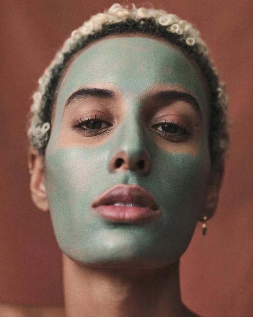 Close-up portrait of a young adult female with a green facial mask, fair skin, and light hair against a muted reddish-brown background.