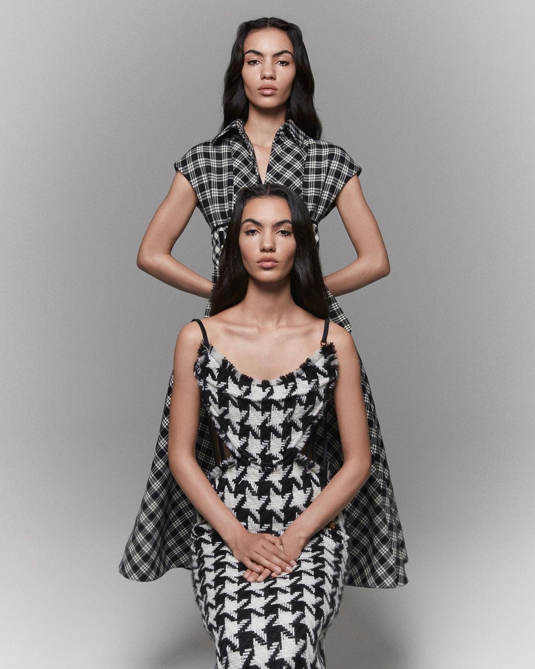 Two women pose in black-and-white patterned dresses against a grey backdrop in a fashion-forward portrait showcasing unique designs.