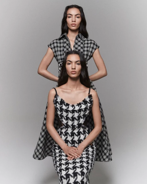 Two women pose in black-and-white patterned dresses against a grey backdrop in a fashion-forward portrait showcasing unique designs.