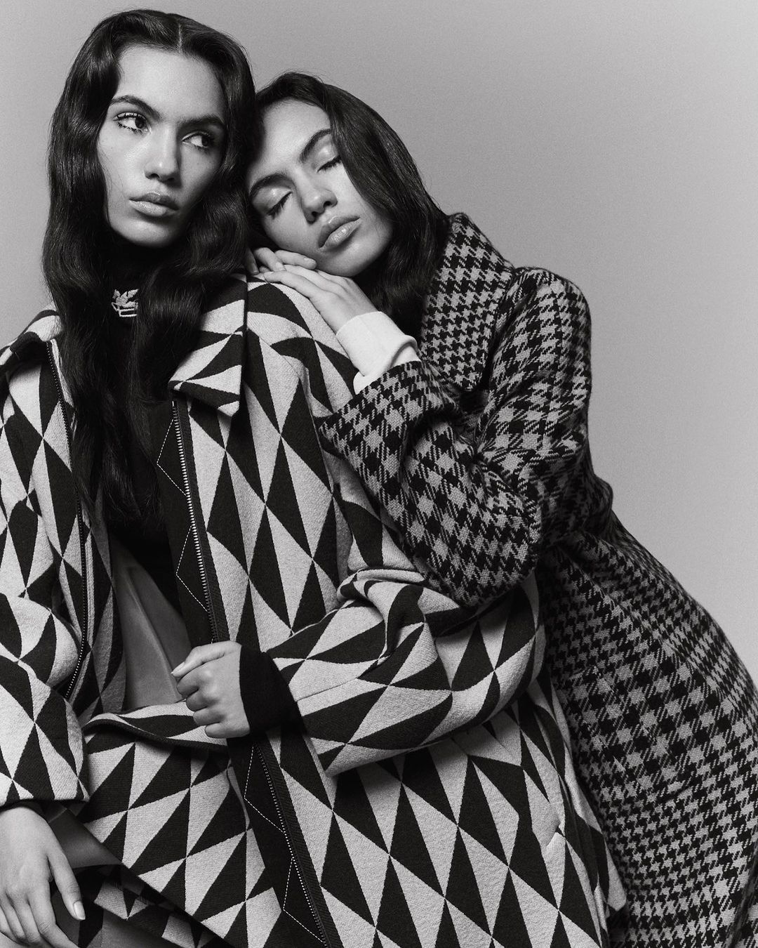 Two women in patterned coats pose in a studio. One woman leans on the other, who looks away. The image is black and white.