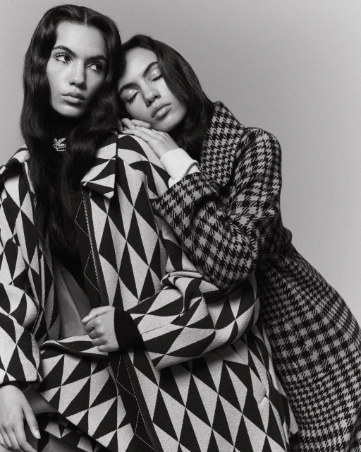 Two women in patterned coats pose in a studio. One woman leans on the other, who looks away. The image is black and white.