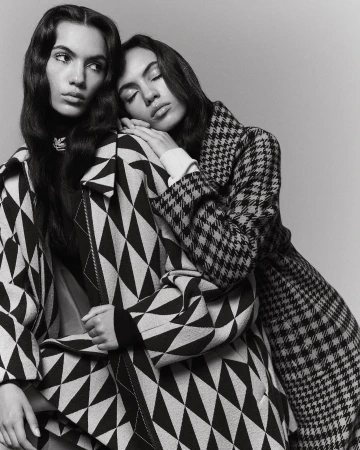 Two women in patterned coats pose in a studio. One woman leans on the other, who looks away. The image is black and white.