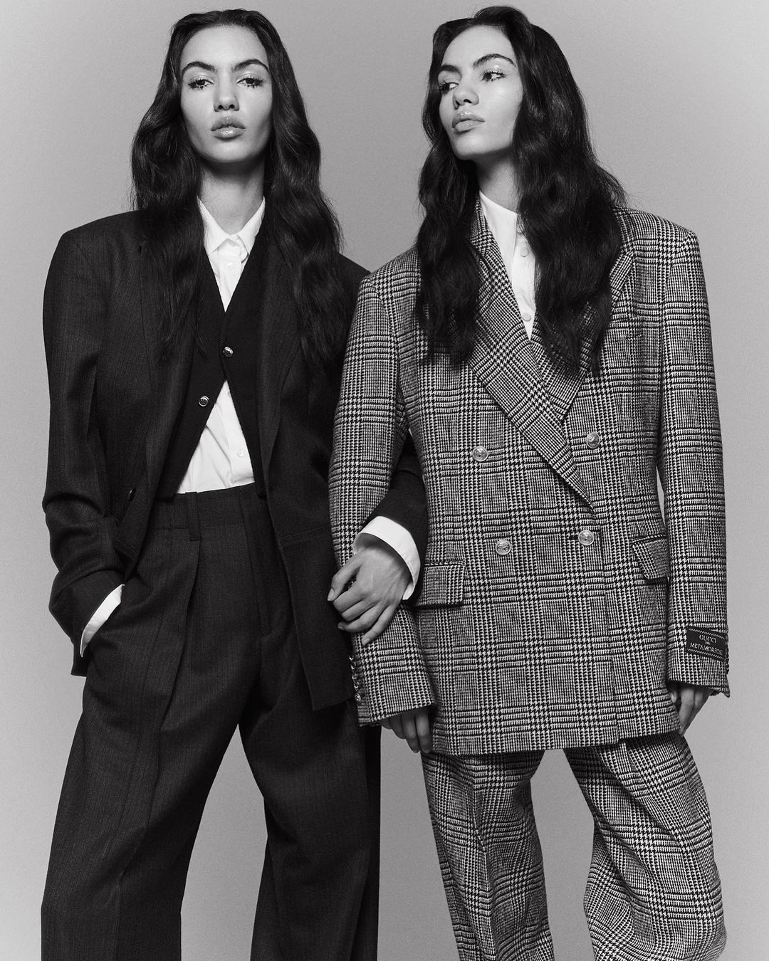 Two women in oversized suits, holding hands, in a black and white studio fashion shot, showcasing tailored blazers and wide-leg trousers.