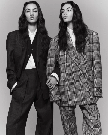 Two women in oversized suits, holding hands, in a black and white studio fashion shot, showcasing tailored blazers and wide-leg trousers.