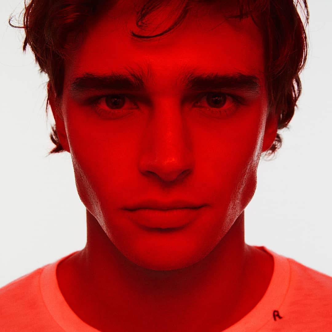 Portrait of a young adult man with dark hair and a white shirt, illuminated in vivid red lighting against a neutral background.