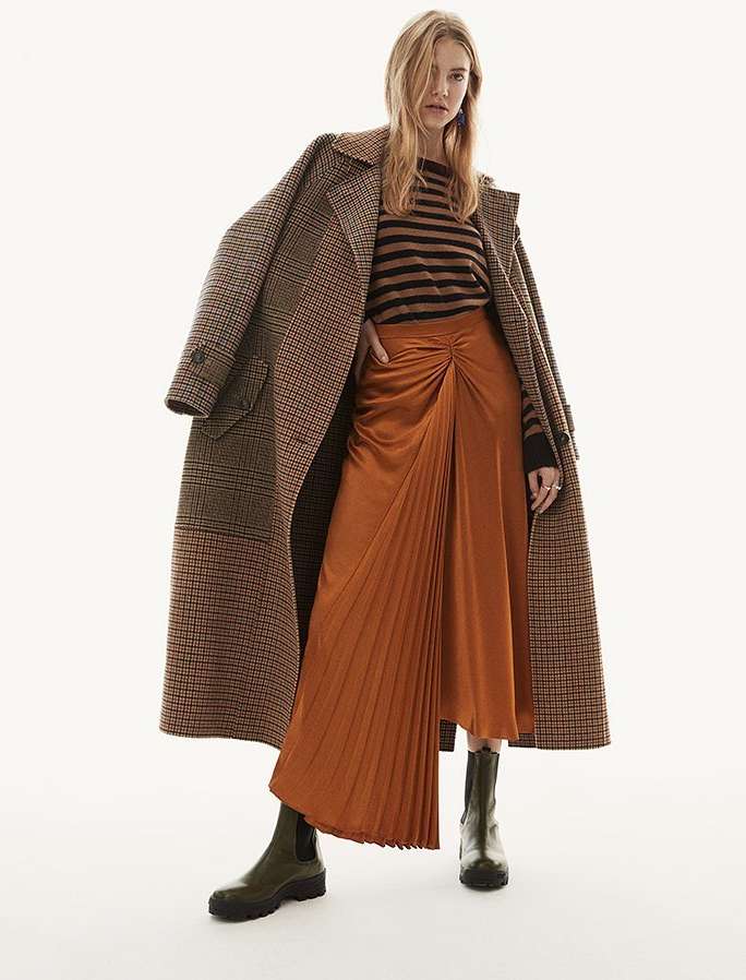 Model wearing a houndstooth coat draped over shoulders, striped sweater, and pleated orange skirt against a white background