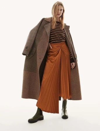 Model wearing a houndstooth coat draped over shoulders, striped sweater, and pleated orange skirt against a white background