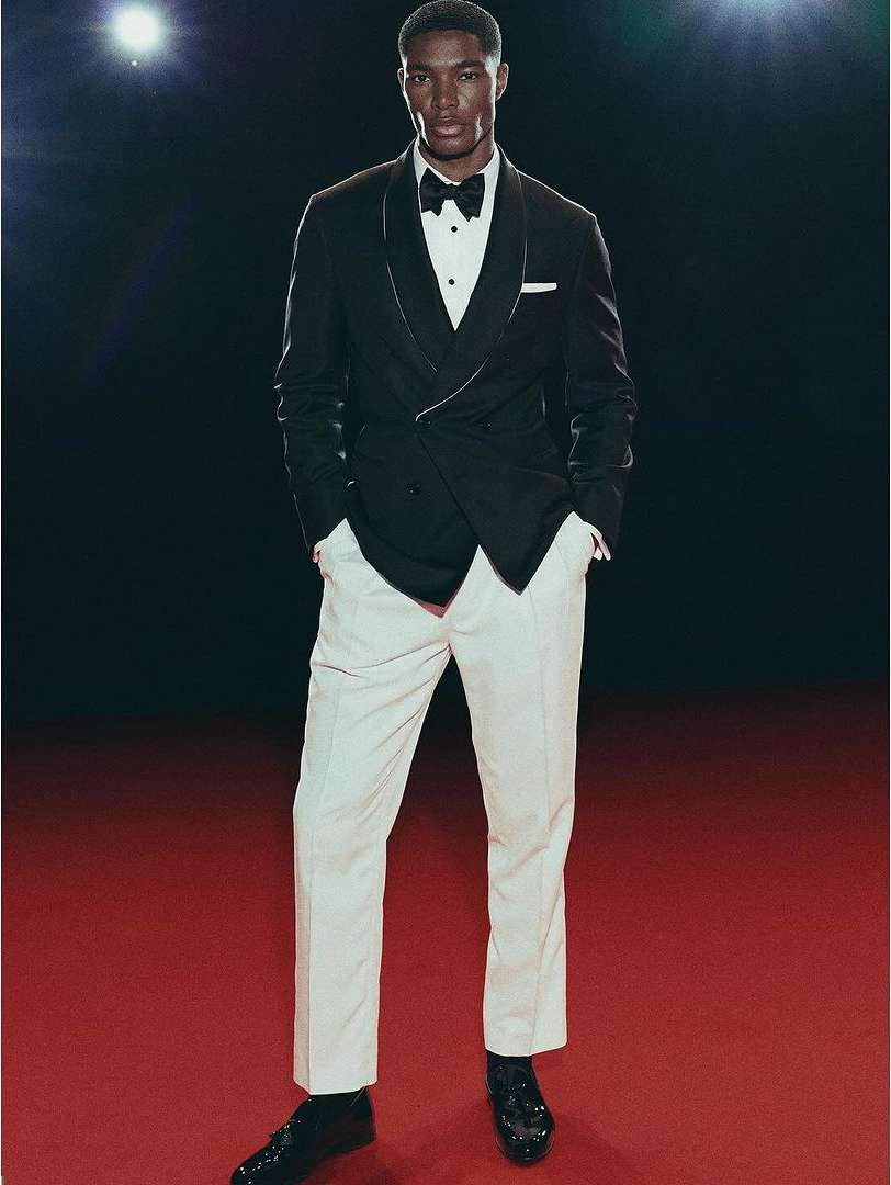 Full-length studio shot of an adult male fashion model in a classic black tuxedo with a bow tie, white dress shirt, and cream-colored pants.