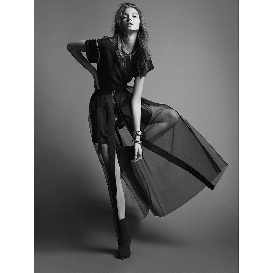 Fashionable woman poses in a dark T-shirt and sheer maxi skirt against a gray background in a studio shot, creating a bold, stylish portrait.
