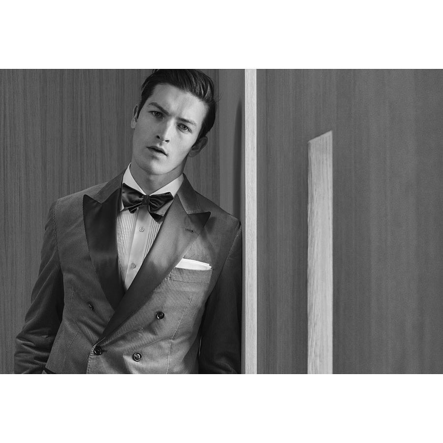 Portrait of a dark-haired adult male wearing a suit, white shirt, and bow tie, leaning against a modern paneled wall in a photo studio.