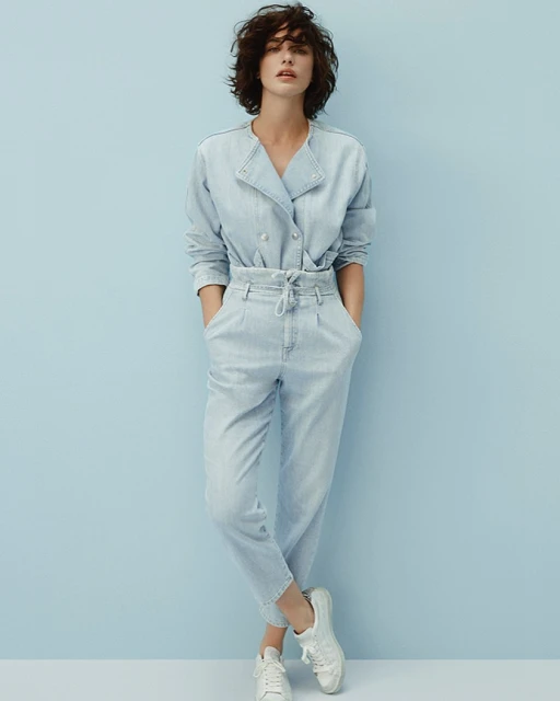 Full shot of an adult woman posing in a light-blue denim double-breasted jacket and matching denim trousers, paired with white sneakers.