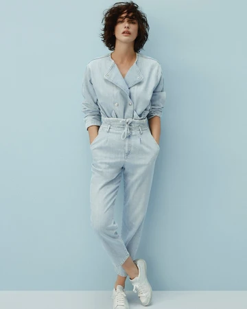 Full shot of an adult woman posing in a light-blue denim double-breasted jacket and matching denim trousers, paired with white sneakers.