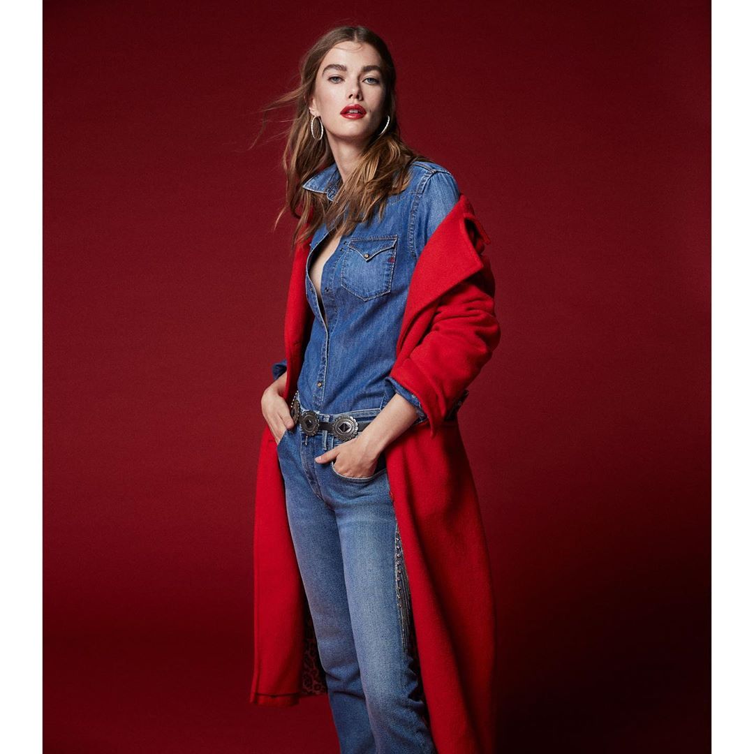 An adult woman poses in a denim outfit and a red coat in a full shot. She is standing against a plain red background.