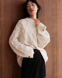 Asian woman wearing an oversized white cable-knit sweater leaning against a warm wood panel wall