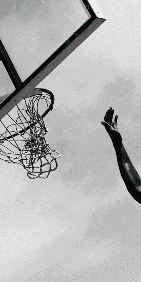 Monochrome upward shot of a basketball hoop, net, backboard, and an extended arm reaching towards the hoop against a cloudy sky.