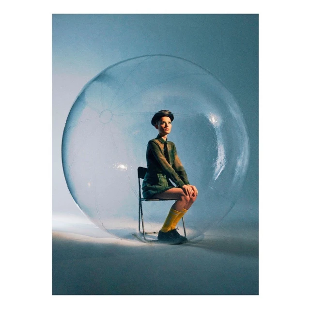 Woman sits inside a large transparent bubble wearing a green suit, black hat, and yellow socks in a studio with soft blue and white lighting.