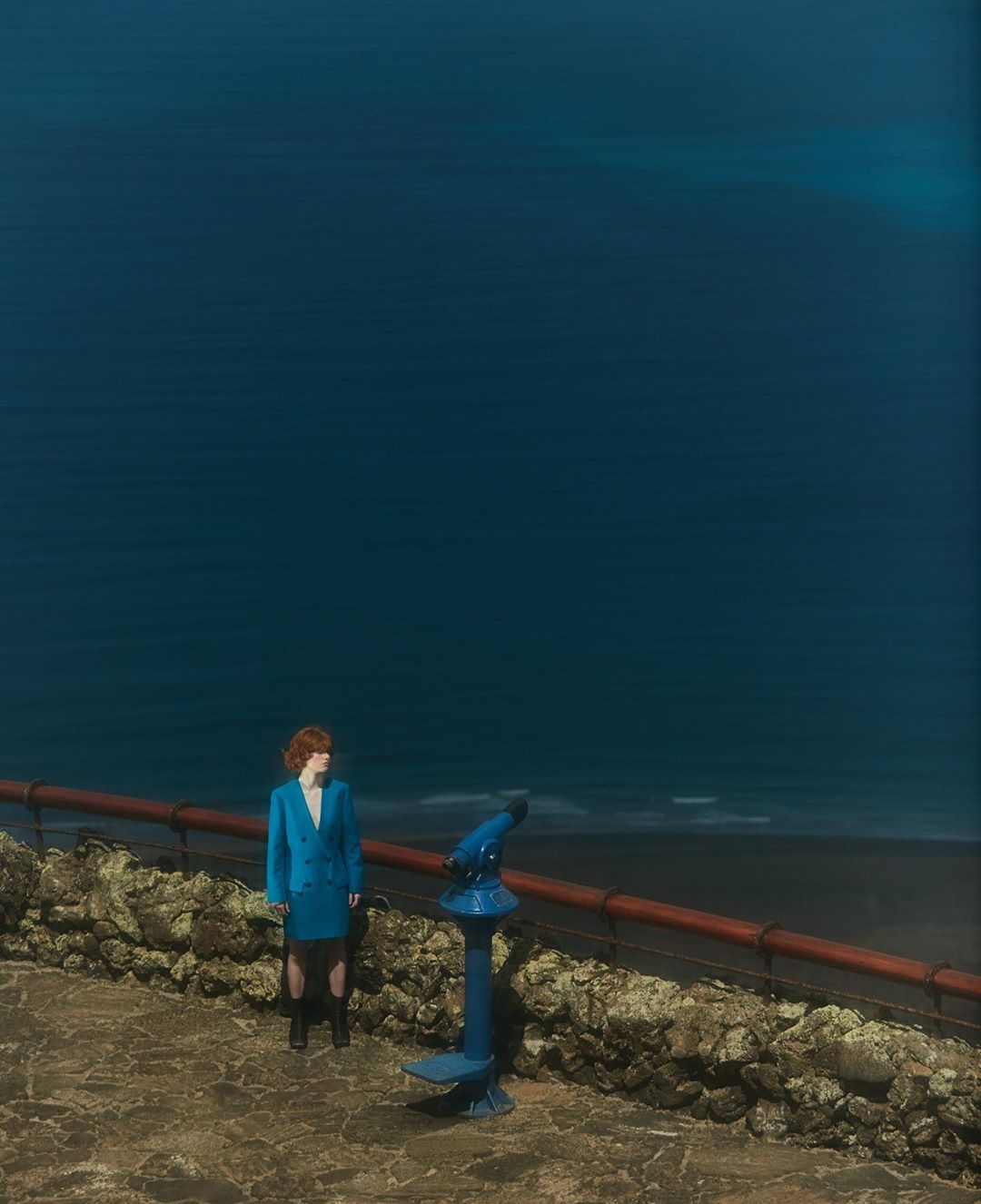 Woman in blue suit stands near a telescope overlooking a dark blue sea from a stone walkway with a stone wall and red railing.