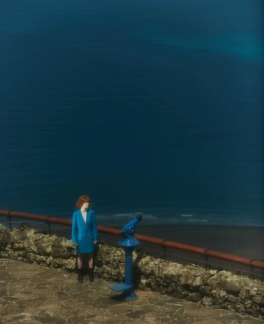 Woman in blue suit stands near a telescope overlooking a dark blue sea from a stone walkway with a stone wall and red railing.
