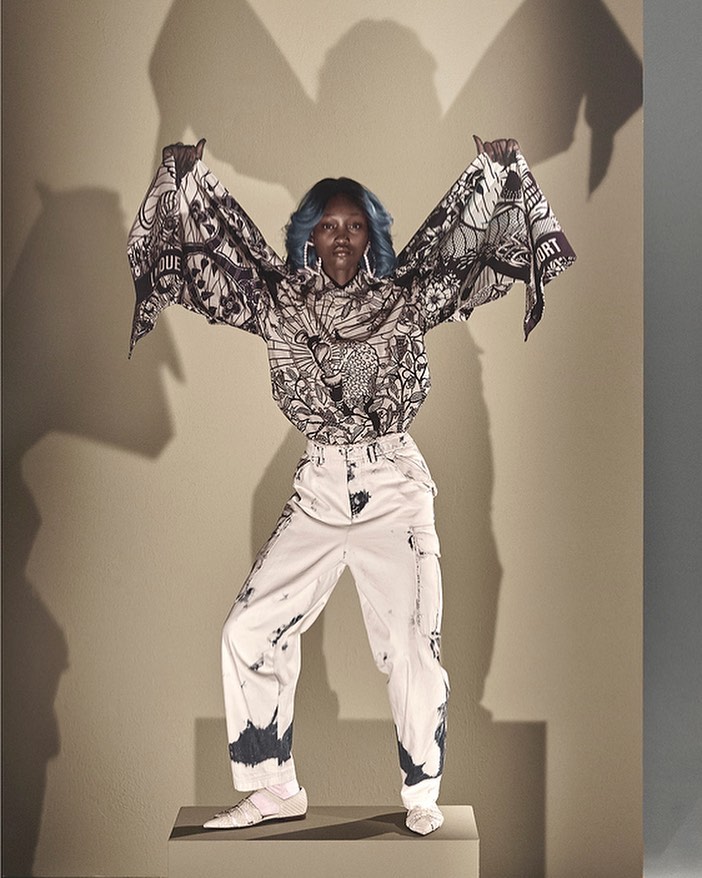 Full-length studio shot of a Black woman in a patterned blouse and white pants standing on a gray block with a gray background.