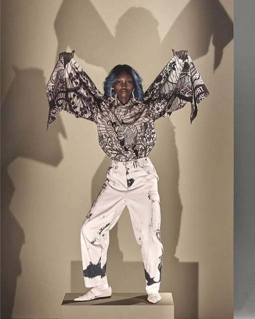 Full-length studio shot of a Black woman in a patterned blouse and white pants standing on a gray block with a gray background.