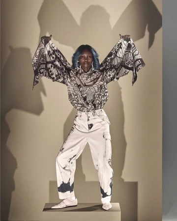 Full-length studio shot of a Black woman in a patterned blouse and white pants standing on a gray block with a gray background.