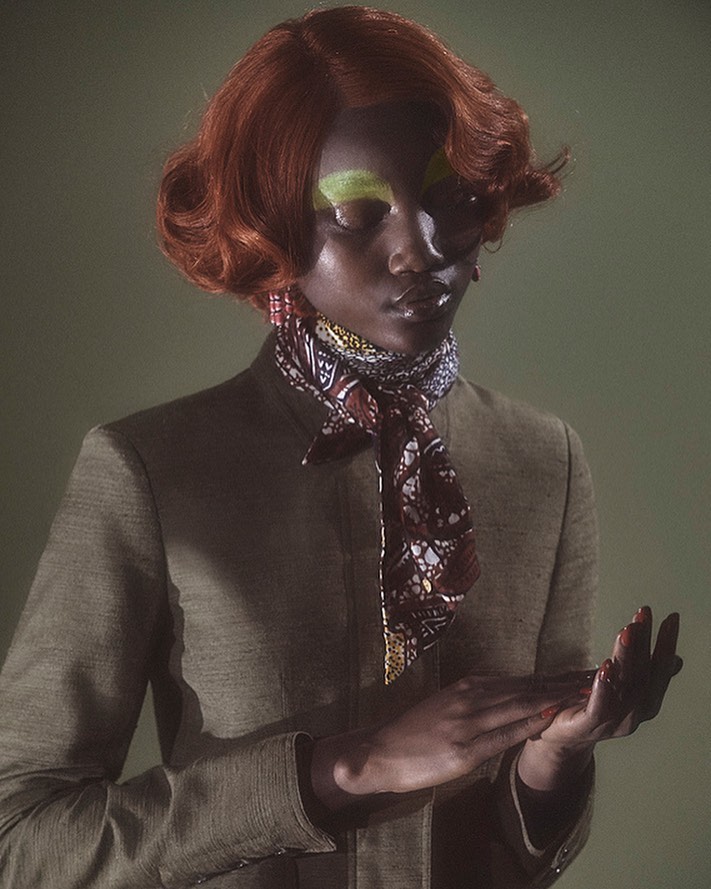 Dark-skinned female model with auburn wavy hair wearing a blazer, patterned scarf, and vibrant green eyeshadow in a studio setting.