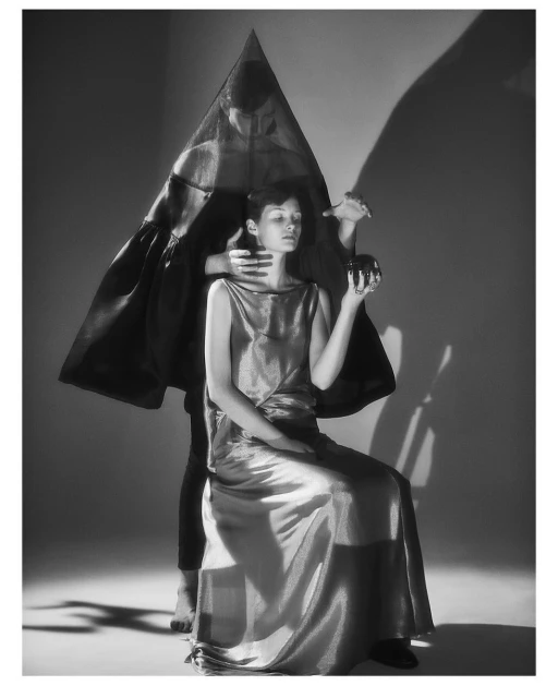 Monochromatic fashion portrait of a woman seated in a gown with another figure standing behind her, holding an orb, in a studio setting.