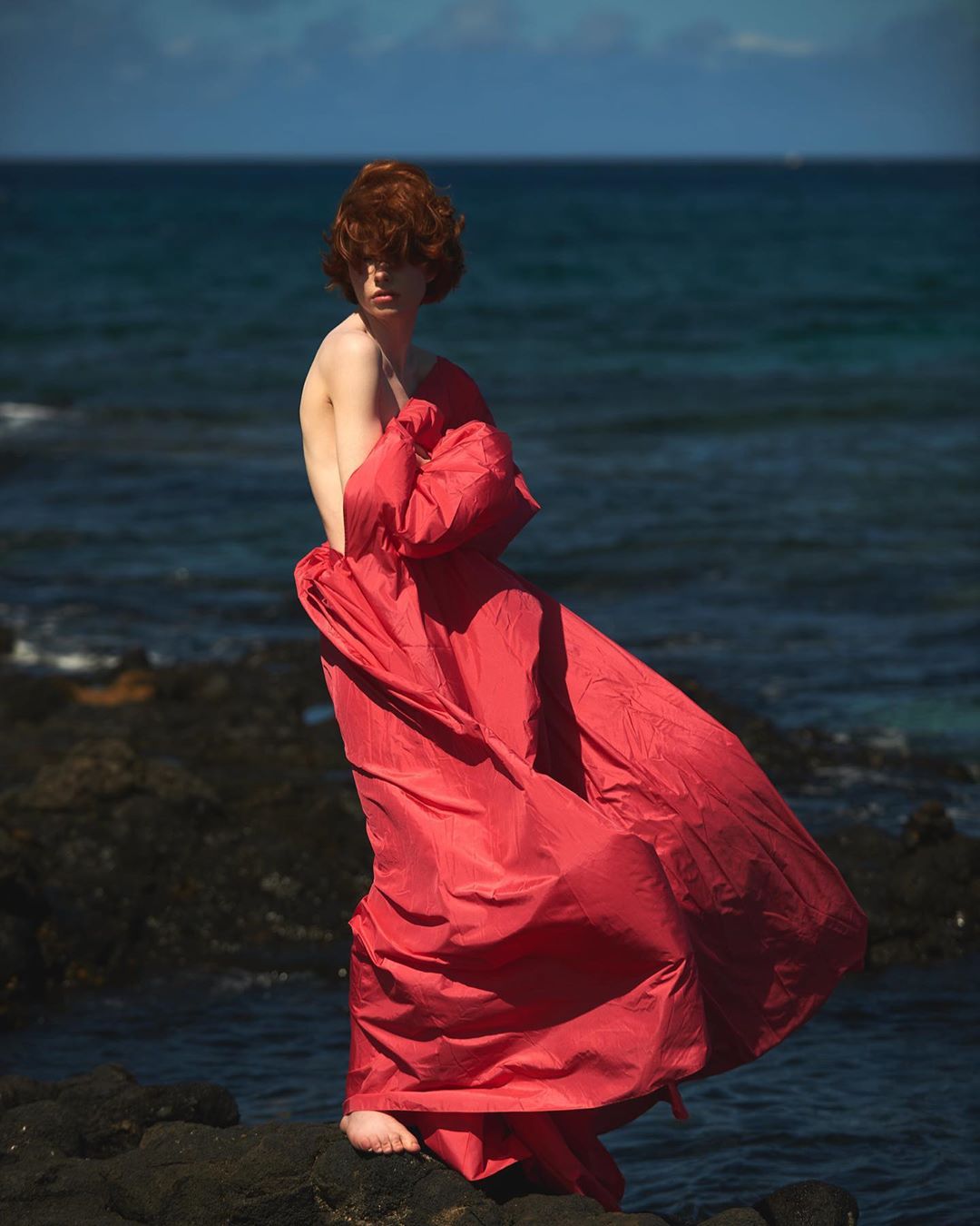 Barefoot person with red hair stands on rocky shore draped in a red cloth, ocean backdrop, under daylight. A vibrant coastal scene.