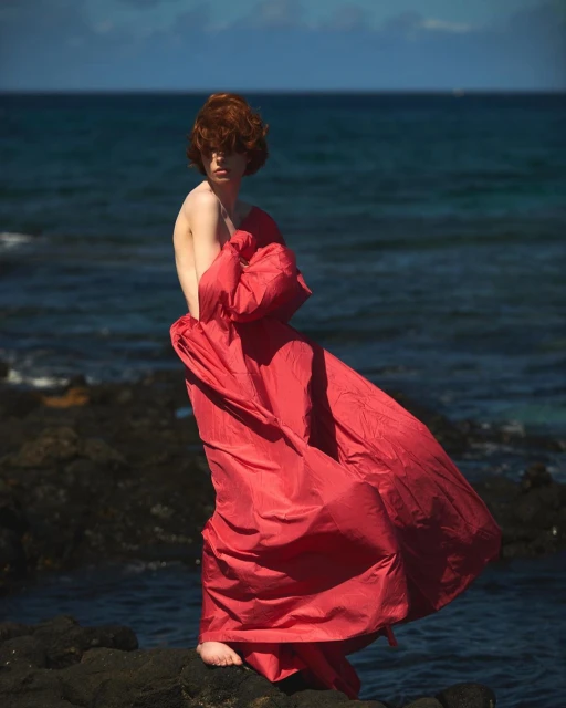 Barefoot person with red hair stands on rocky shore draped in a red cloth, ocean backdrop, under daylight. A vibrant coastal scene.