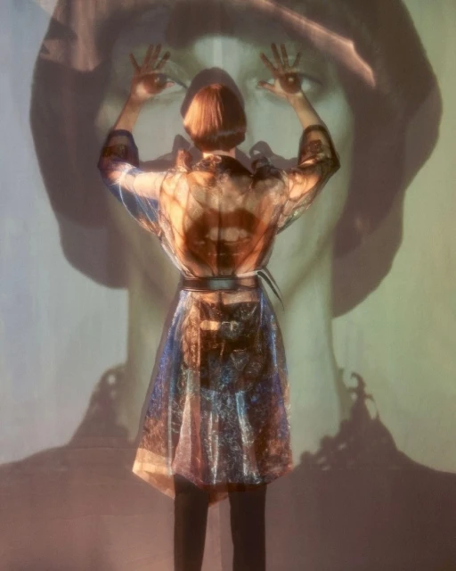 Woman in transparent coat faces projected face with hands up. Back view of adult with short red hair in an indoor environment.