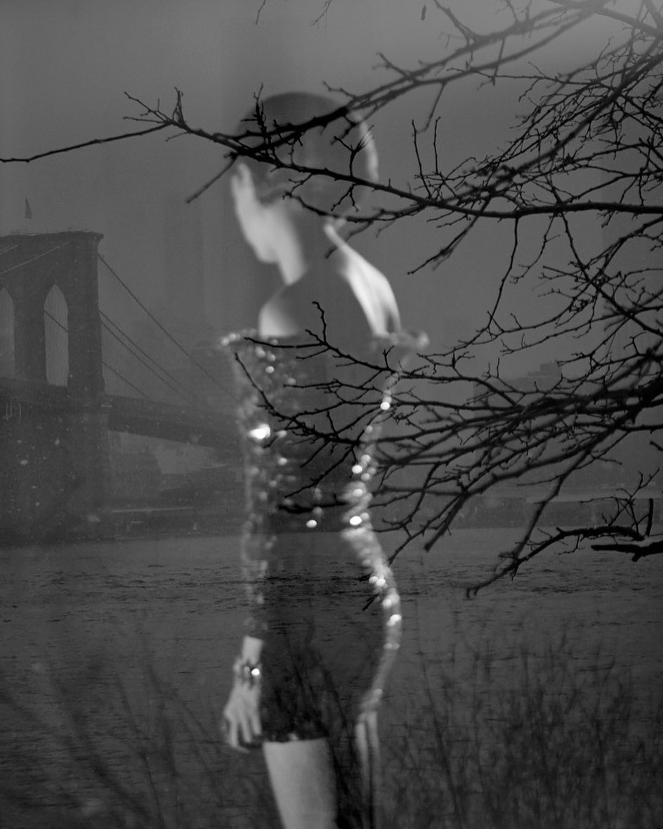 Black and white image of a woman in a sequin dress near the Brooklyn Bridge, framed by bare tree branches, evoking a moody, layered atmosphere.