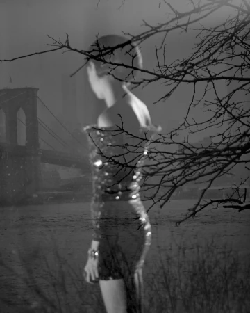 Black and white image of a woman in a sequin dress near the Brooklyn Bridge, framed by bare tree branches, evoking a moody, layered atmosphere.