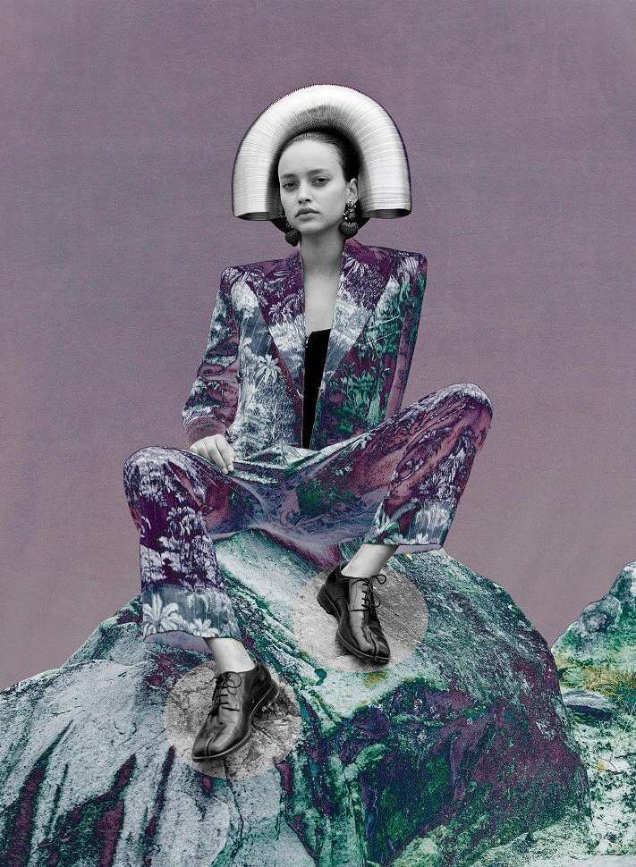 A woman sits on a rock wearing a floral-patterned suit and a large metal headdress. She has a neutral expression and is looking forward.