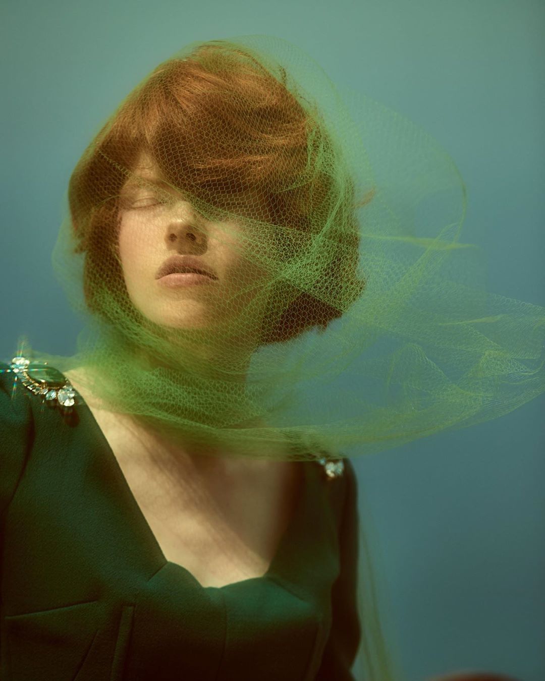 Woman with red hair and closed eyes, covered by green veil, wearing dark green dress with jeweled epaulettes against a blue backdrop.