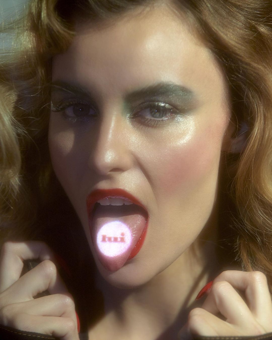 Close-up of a young adult woman with curly hair sticking out her tongue, which has text on it, glossy makeup, and red lipstick.